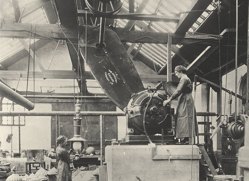 Black-and-white photograph of a workshop under a glass roof. A woman stands on a platform beside a large electric motor. A long chain-drive casing marked “The Coventry Chain Company Ltd” rises from it to a pulley on the shaft under the roof. Another woman works at a bench below.
